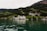 A picturesque lakeside scene features a large, elegant building with a white facade situated among lush green hills. Surrounding the main structure are smaller buildings, with a backdrop of dense, green forested mountains. The lake in the foreground has clear greenish water reflecting the scenic landscape.