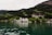 A picturesque lakeside scene features a large, elegant building with a white facade situated among lush green hills. Surrounding the main structure are smaller buildings, with a backdrop of dense, green forested mountains. The lake in the foreground has clear greenish water reflecting the scenic landscape.