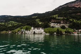 A picturesque lakeside scene features a large, elegant building with a white facade situated among lush green hills. Surrounding the main structure are smaller buildings, with a backdrop of dense, green forested mountains. The lake in the foreground has clear greenish water reflecting the scenic landscape.