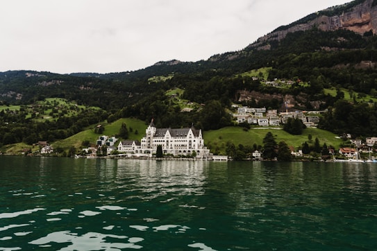A picturesque lakeside scene features a large, elegant building with a white facade situated among lush green hills. Surrounding the main structure are smaller buildings, with a backdrop of dense, green forested mountains. The lake in the foreground has clear greenish water reflecting the scenic landscape.