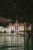 A grand, traditional hotel with intricate red and white detailing is set against a lush, forested mountain backdrop. The hotel has a steep, pointed roof and a distinct tower. It sits at the edge of a serene body of water, reflecting the building and surrounding greenery.