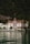 A grand, traditional hotel with intricate red and white detailing is set against a lush, forested mountain backdrop. The hotel has a steep, pointed roof and a distinct tower. It sits at the edge of a serene body of water, reflecting the building and surrounding greenery.