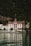 A grand, traditional hotel with intricate red and white detailing is set against a lush, forested mountain backdrop. The hotel has a steep, pointed roof and a distinct tower. It sits at the edge of a serene body of water, reflecting the building and surrounding greenery.