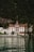 A grand, traditional hotel with intricate red and white detailing is set against a lush, forested mountain backdrop. The hotel has a steep, pointed roof and a distinct tower. It sits at the edge of a serene body of water, reflecting the building and surrounding greenery.