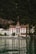 A grand, traditional hotel with intricate red and white detailing is set against a lush, forested mountain backdrop. The hotel has a steep, pointed roof and a distinct tower. It sits at the edge of a serene body of water, reflecting the building and surrounding greenery.