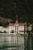 A grand, traditional hotel with intricate red and white detailing is set against a lush, forested mountain backdrop. The hotel has a steep, pointed roof and a distinct tower. It sits at the edge of a serene body of water, reflecting the building and surrounding greenery.
