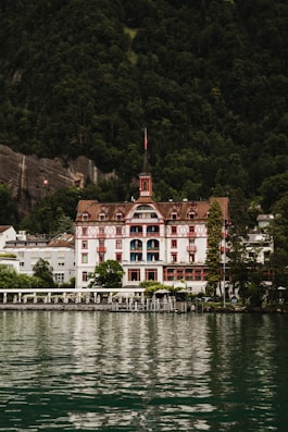 A grand, traditional hotel with intricate red and white detailing is set against a lush, forested mountain backdrop. The hotel has a steep, pointed roof and a distinct tower. It sits at the edge of a serene body of water, reflecting the building and surrounding greenery.