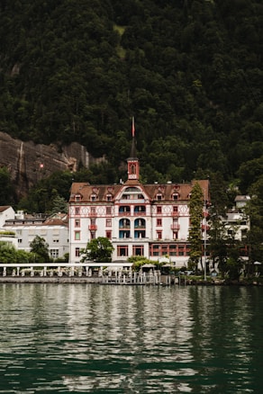 A grand, traditional hotel with intricate red and white detailing is set against a lush, forested mountain backdrop. The hotel has a steep, pointed roof and a distinct tower. It sits at the edge of a serene body of water, reflecting the building and surrounding greenery.