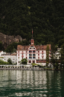 A grand, traditional hotel with intricate red and white detailing is set against a lush, forested mountain backdrop. The hotel has a steep, pointed roof and a distinct tower. It sits at the edge of a serene body of water, reflecting the building and surrounding greenery.
