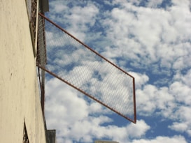 A metal frame with a mesh netting extends outward from a building's surface, set against a backdrop of a sky filled with scattered, fluffy white clouds.