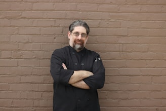 A person with glasses and a beard stands confidently with arms crossed, wearing a black chef's coat, against a textured brown brick wall.