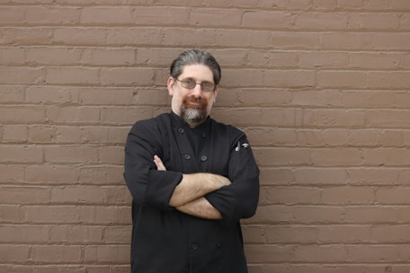 A person with glasses and a beard stands confidently with arms crossed, wearing a black chef's coat, against a textured brown brick wall.