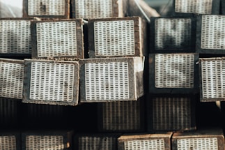 A stack of rectangular wooden beams with white perforated metal plates on one end, arranged in an orderly manner. The beams have a weathered appearance, and the lighting suggests a natural, possibly outdoor setting.