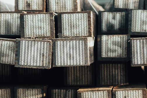 A stack of rectangular wooden beams with white perforated metal plates on one end, arranged in an orderly manner. The beams have a weathered appearance, and the lighting suggests a natural, possibly outdoor setting.