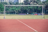 A soccer field with two visible goalposts is surrounded by lush green trees. In the background, tall buildings can be seen, indicating an urban environment. The field comprises an empty grass area, with a running track of reddish-brown color surrounding it.