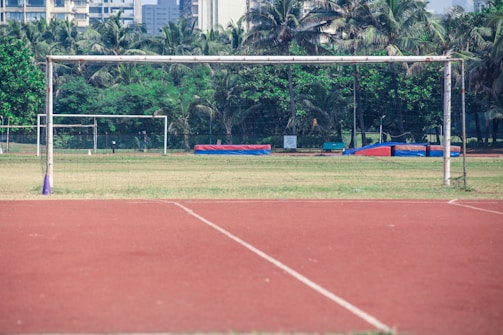 A soccer field with two visible goalposts is surrounded by lush green trees. In the background, tall buildings can be seen, indicating an urban environment. The field comprises an empty grass area, with a running track of reddish-brown color surrounding it.