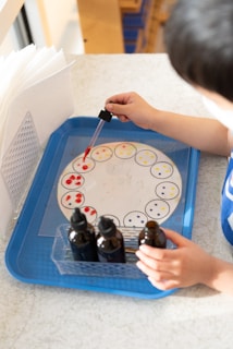 A child is using a dropper to add colored liquid to a circular paper with sections, presumably for an experiment or art project. There are several brown bottles and a blue tray involved, along with paper towels nearby.