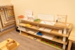 A neatly organized classroom shelf contains a variety of educational materials and tools. There are wooden trays, colored boxes, abacuses, and charts. Each shelf is filled with learning aids, prominently featuring items like math boards and counting tools. The environment is bright and has a warm, inviting atmosphere typical for a Montessori setting.