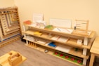 A neatly organized classroom shelf contains a variety of educational materials and tools. There are wooden trays, colored boxes, abacuses, and charts. Each shelf is filled with learning aids, prominently featuring items like math boards and counting tools. The environment is bright and has a warm, inviting atmosphere typical for a Montessori setting.