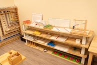 A neatly organized classroom shelf contains a variety of educational materials and tools. There are wooden trays, colored boxes, abacuses, and charts. Each shelf is filled with learning aids, prominently featuring items like math boards and counting tools. The environment is bright and has a warm, inviting atmosphere typical for a Montessori setting.