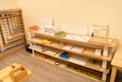 A neatly organized classroom shelf contains a variety of educational materials and tools. There are wooden trays, colored boxes, abacuses, and charts. Each shelf is filled with learning aids, prominently featuring items like math boards and counting tools. The environment is bright and has a warm, inviting atmosphere typical for a Montessori setting.