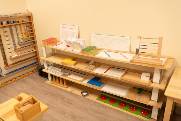 A neatly organized classroom shelf contains a variety of educational materials and tools. There are wooden trays, colored boxes, abacuses, and charts. Each shelf is filled with learning aids, prominently featuring items like math boards and counting tools. The environment is bright and has a warm, inviting atmosphere typical for a Montessori setting.