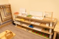 A neatly organized classroom shelf contains a variety of educational materials and tools. There are wooden trays, colored boxes, abacuses, and charts. Each shelf is filled with learning aids, prominently featuring items like math boards and counting tools. The environment is bright and has a warm, inviting atmosphere typical for a Montessori setting.