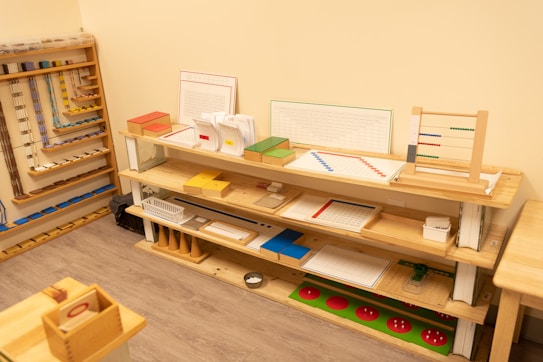 A neatly organized classroom shelf contains a variety of educational materials and tools. There are wooden trays, colored boxes, abacuses, and charts. Each shelf is filled with learning aids, prominently featuring items like math boards and counting tools. The environment is bright and has a warm, inviting atmosphere typical for a Montessori setting.