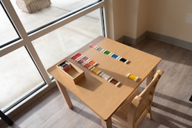 A wooden table is placed near a window, with colored tiles arranged neatly in a pattern and a small box holding additional tiles. There is a wooden chair next to the table, and the floor is made of light-colored wood.