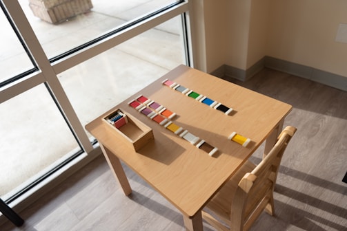 A wooden table is placed near a window, with colored tiles arranged neatly in a pattern and a small box holding additional tiles. There is a wooden chair next to the table, and the floor is made of light-colored wood.