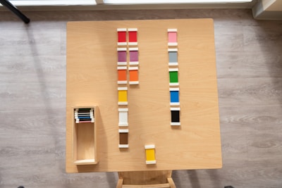 Wooden table with neatly arranged colorful tiles in various shades. The tiles are lined up in rows, and there is a wooden box on the left containing additional tiles.