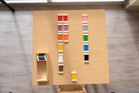 Wooden table with neatly arranged colorful tiles in various shades. The tiles are lined up in rows, and there is a wooden box on the left containing additional tiles.