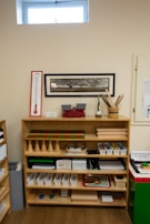 A wooden shelf filled with educational materials is set against a beige wall. The shelf is organized with a variety of items such as wooden cones, trays, baskets, and plastic bins. Above the shelf, a rectangular, framed image of a tree is mounted on the wall. There is a thermometer chart to the left side and a small window near the ceiling allowing natural light in.