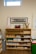 A wooden shelf filled with educational materials is set against a beige wall. The shelf is organized with a variety of items such as wooden cones, trays, baskets, and plastic bins. Above the shelf, a rectangular, framed image of a tree is mounted on the wall. There is a thermometer chart to the left side and a small window near the ceiling allowing natural light in.