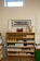 A wooden shelf filled with educational materials is set against a beige wall. The shelf is organized with a variety of items such as wooden cones, trays, baskets, and plastic bins. Above the shelf, a rectangular, framed image of a tree is mounted on the wall. There is a thermometer chart to the left side and a small window near the ceiling allowing natural light in.
