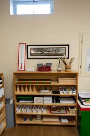 A wooden shelf filled with educational materials is set against a beige wall. The shelf is organized with a variety of items such as wooden cones, trays, baskets, and plastic bins. Above the shelf, a rectangular, framed image of a tree is mounted on the wall. There is a thermometer chart to the left side and a small window near the ceiling allowing natural light in.