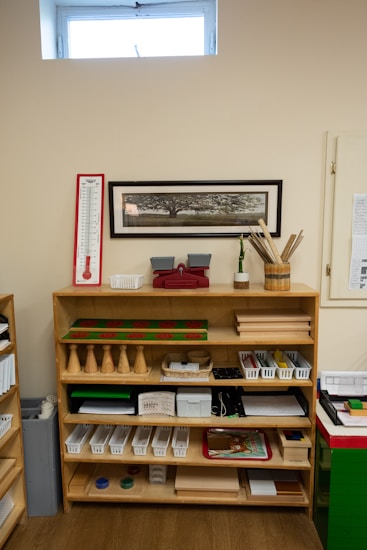 A wooden shelf filled with educational materials is set against a beige wall. The shelf is organized with a variety of items such as wooden cones, trays, baskets, and plastic bins. Above the shelf, a rectangular, framed image of a tree is mounted on the wall. There is a thermometer chart to the left side and a small window near the ceiling allowing natural light in.