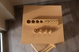 A wooden cylinder block puzzle is placed on a square wooden table, seen from above. The puzzle consists of a rectangular block with various cylindrical holes and corresponding wooden pegs of different sizes placed nearby. The surface is illuminated by warm light, casting soft shadows on the table.
