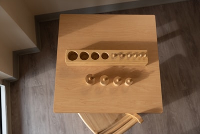 A wooden cylinder block puzzle is placed on a square wooden table, seen from above. The puzzle consists of a rectangular block with various cylindrical holes and corresponding wooden pegs of different sizes placed nearby. The surface is illuminated by warm light, casting soft shadows on the table.