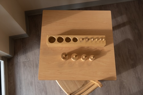 A wooden cylinder block puzzle is placed on a square wooden table, seen from above. The puzzle consists of a rectangular block with various cylindrical holes and corresponding wooden pegs of different sizes placed nearby. The surface is illuminated by warm light, casting soft shadows on the table.
