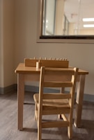 A small wooden table and chair set placed against a wall, with a window partially visible behind it. On top of the table, there is a wooden educational toy with small pegs, possibly for sorting or counting.