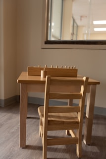 A small wooden table and chair set placed against a wall, with a window partially visible behind it. On top of the table, there is a wooden educational toy with small pegs, possibly for sorting or counting.