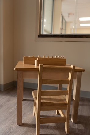 A small wooden table and chair set placed against a wall, with a window partially visible behind it. On top of the table, there is a wooden educational toy with small pegs, possibly for sorting or counting.