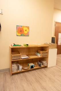 A light-colored wooden shelf is organized with various educational materials. The top shelf holds trays with papers and a small green basket. The middle shelf has more paper trays and a small container. The bottom shelf contains baskets, small books, and plastic folders. A vibrant painting of colorful flowers is hung above the shelf on a cream-colored wall. The floor is wooden, and there is a small chalkboard next to the shelf.