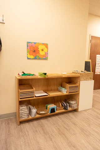 A light-colored wooden shelf is organized with various educational materials. The top shelf holds trays with papers and a small green basket. The middle shelf has more paper trays and a small container. The bottom shelf contains baskets, small books, and plastic folders. A vibrant painting of colorful flowers is hung above the shelf on a cream-colored wall. The floor is wooden, and there is a small chalkboard next to the shelf.