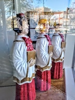 Three choir figurines dressed in red robes with white and gold details are displayed behind a shop window. Each holds a songbook and faces the same direction, with reflections of a street scene visible in the glass. The reflection shows buildings, trees, and a bright blue sky.