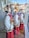 Three choir figurines dressed in red robes with white and gold details are displayed behind a shop window. Each holds a songbook and faces the same direction, with reflections of a street scene visible in the glass. The reflection shows buildings, trees, and a bright blue sky.