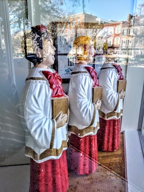 Three choir figurines dressed in red robes with white and gold details are displayed behind a shop window. Each holds a songbook and faces the same direction, with reflections of a street scene visible in the glass. The reflection shows buildings, trees, and a bright blue sky.