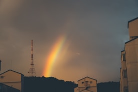 A vibrant rainbow arcs across the sky during an overcast sunset, partially covered by dark clouds. The silhouette of a communication tower stands prominently against the colorful sky, surrounded by several residential buildings in the foreground.