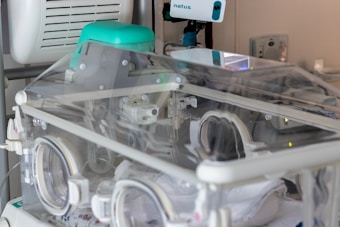 A neonatal incubator with a transparent enclosure, equipped with medical apparatus and monitoring devices. The incubator is designed to provide a controlled environment for newborns requiring medical care.